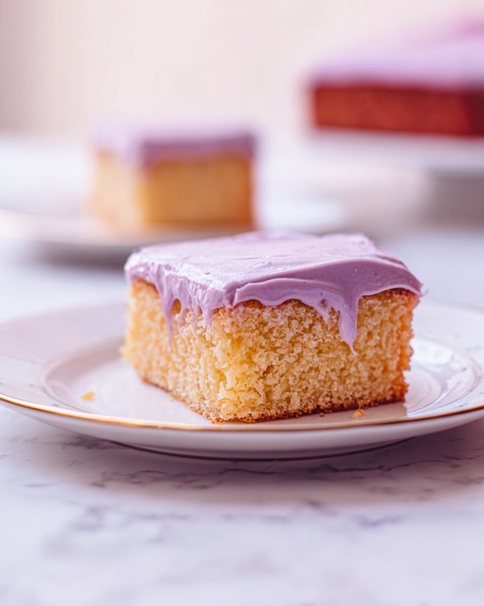 A single square piece of cake sits on a white plate with a thin gold rim, placed on a white marbled surface. The cake has one main layer with a light brown, soft, and fluffy texture that shows small crumbs inside. On top, there is a thick layer of smooth purple frosting that gently drips down the sides of the cake. In the background, a second piece of the same cake is slightly blurred on a matching white plate. Photo taken with an iphone --ar 4:5 --v 7
