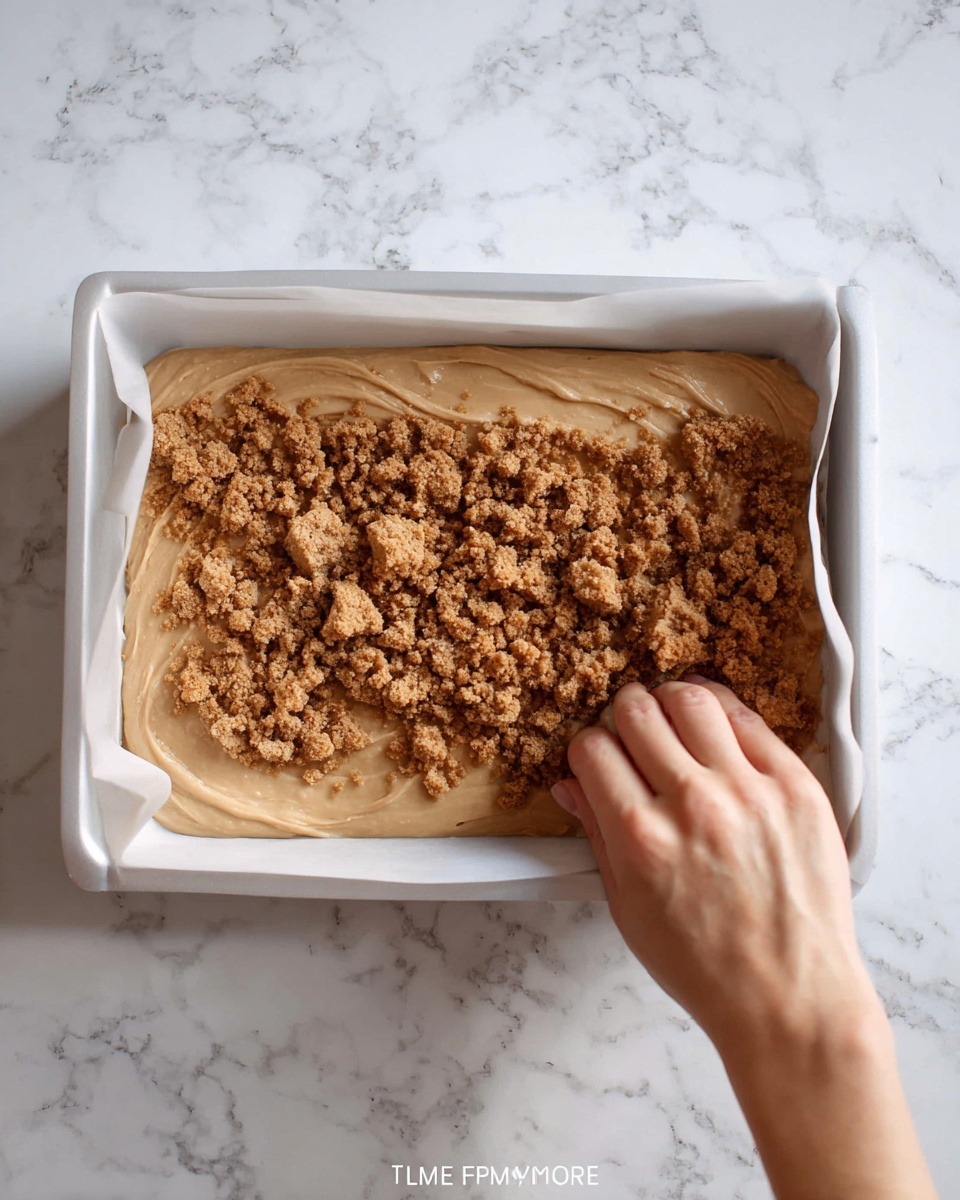 Biscoff Fudge Recipe 4 A white rectangular baking pan lined with parchment paper holds a thick, light tan batter spread evenly inside. On top, crumbly pieces of a darker brown cookie or biscuit are scattered and lightly pressed down with a woman's hand. The scene is set on a white marbled surface. Photo taken with an iphone --ar 4:5 --v 7