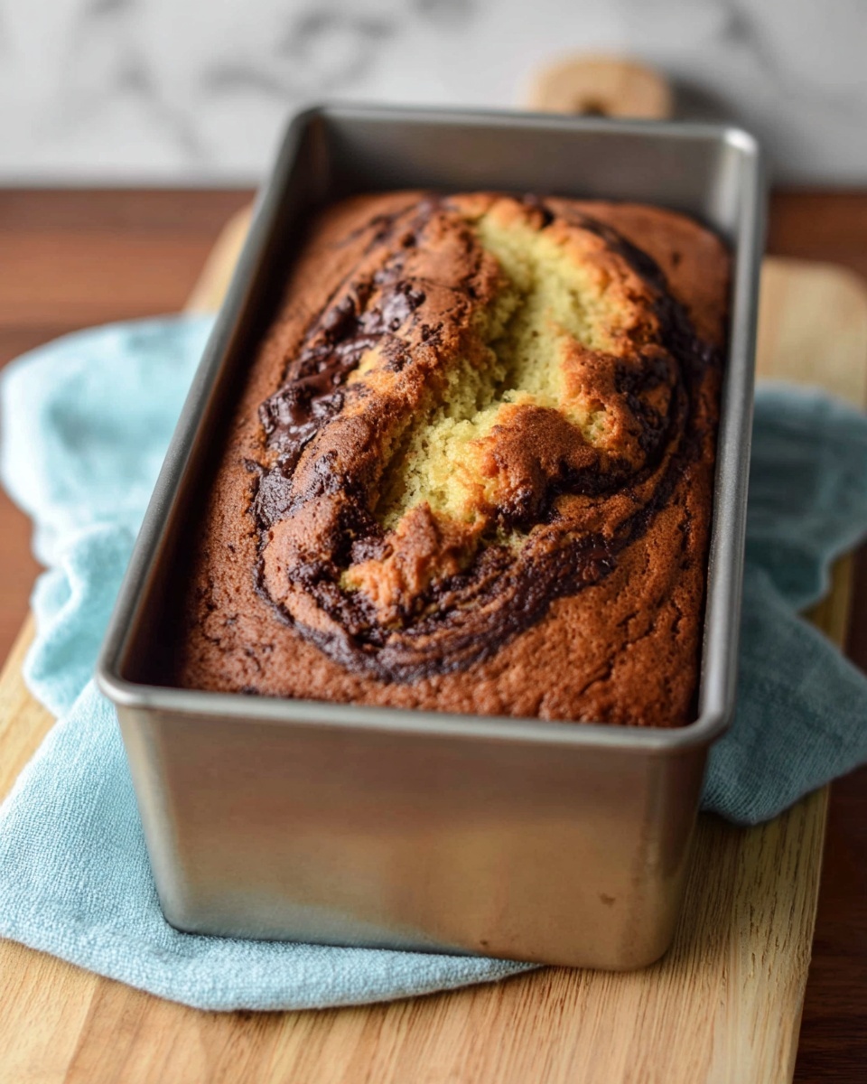 A golden brown loaf cake with dark chocolate swirls on the top, showing a cracked surface revealing a moist inside with light yellow and dark brown marbled layers. The cake sits inside a silver metal loaf pan, which rests on a light blue cloth over a light wooden cutting board. The background has a white marbled texture. Photo taken with an iphone --ar 4:5 --v 7