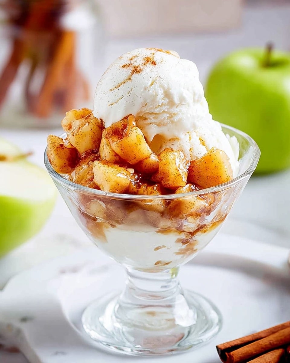 A clear glass dessert bowl holds three scoops of smooth white vanilla ice cream at the bottom layer. On top, there is a generous layer of small, golden-brown cooked apple chunks mixed with cinnamon, giving a slightly shiny and warm look from the syrupy coating. The dessert is set on a white marbled surface, with a blurred background that includes a green apple on the right and a glass jar with cinnamon sticks behind. Photo taken with an iphone --ar 4:5 --v 7