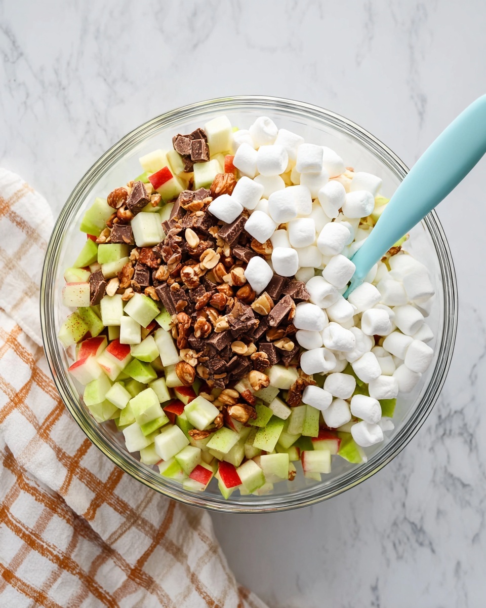 A clear glass bowl sits on a white marbled surface with a checkered cloth beside it. Inside the bowl, the bottom layer is made of small pieces of green and red apple with a firm texture. On top of the fruit layer, there is a second layer of chopped chocolate candy bars, showing a mix of brown chocolate and tan peanut pieces. The top layer consists of a pile of small white marshmallows clustered mostly on one side. A light blue spoon rests inside the bowl against the marshmallows, with the handle pointing outwards. photo taken with an iphone --ar 4:5 --v 7