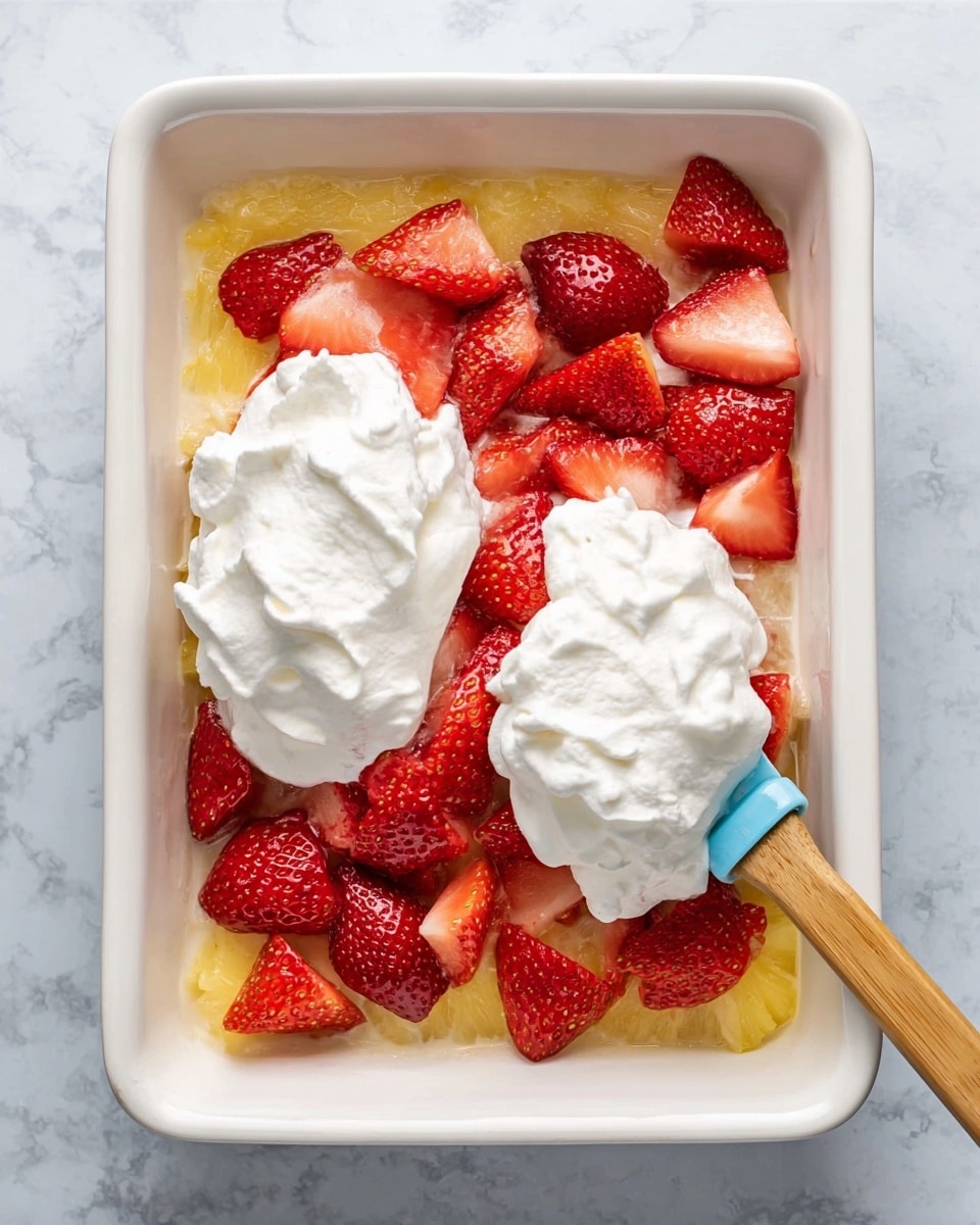 The image shows a white rectangular dish with three visible layers. The bottom layer is crushed pineapple, light yellow with a slightly juicy texture. The middle layer is whole and halved fresh strawberries, bright red with visible seeds and soft texture spread evenly on top of the pineapple. The top layer is two large dollops of white whipped cream placed on the strawberries, one dollop holding a blue spatula with light brown wooden handles, resting slightly on the fruit. The dish is placed on a white marbled surface. Photo taken with an iphone --ar 4:5 --v 7