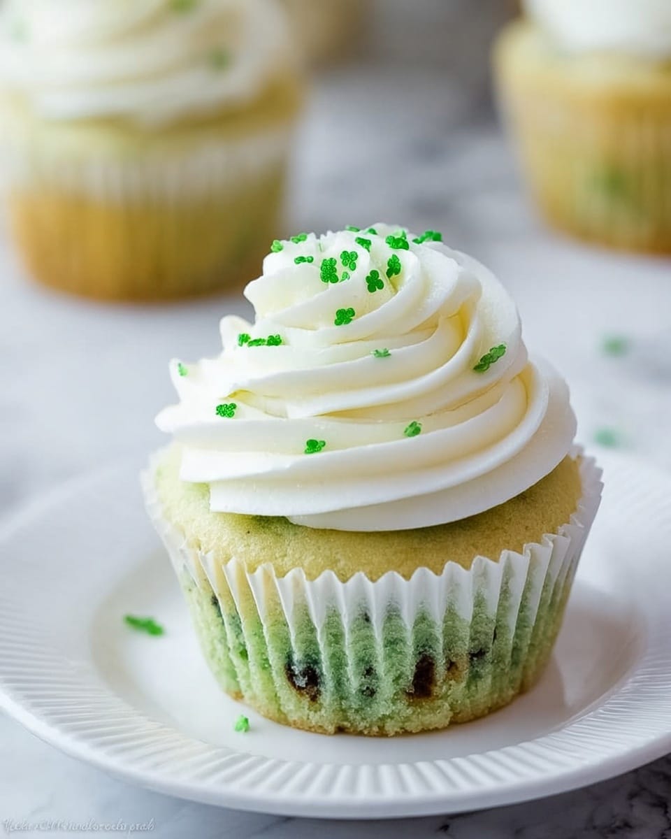 A single cupcake sits on a white plate with subtle ridges around the edge, placed on a white marbled surface. The cupcake has two main layers: the bottom layer is a light green cake with small dark spots inside, wrapped in a white ridged paper liner. On top, there is a thick swirl of smooth white frosting forming a spiral peak. Scattered on the frosting are tiny green clover-shaped sprinkles, adding small bright green details. In the background, a slightly blurred second cupcake with similar colors can be seen. photo taken with an iphone --ar 4:5 --v 7