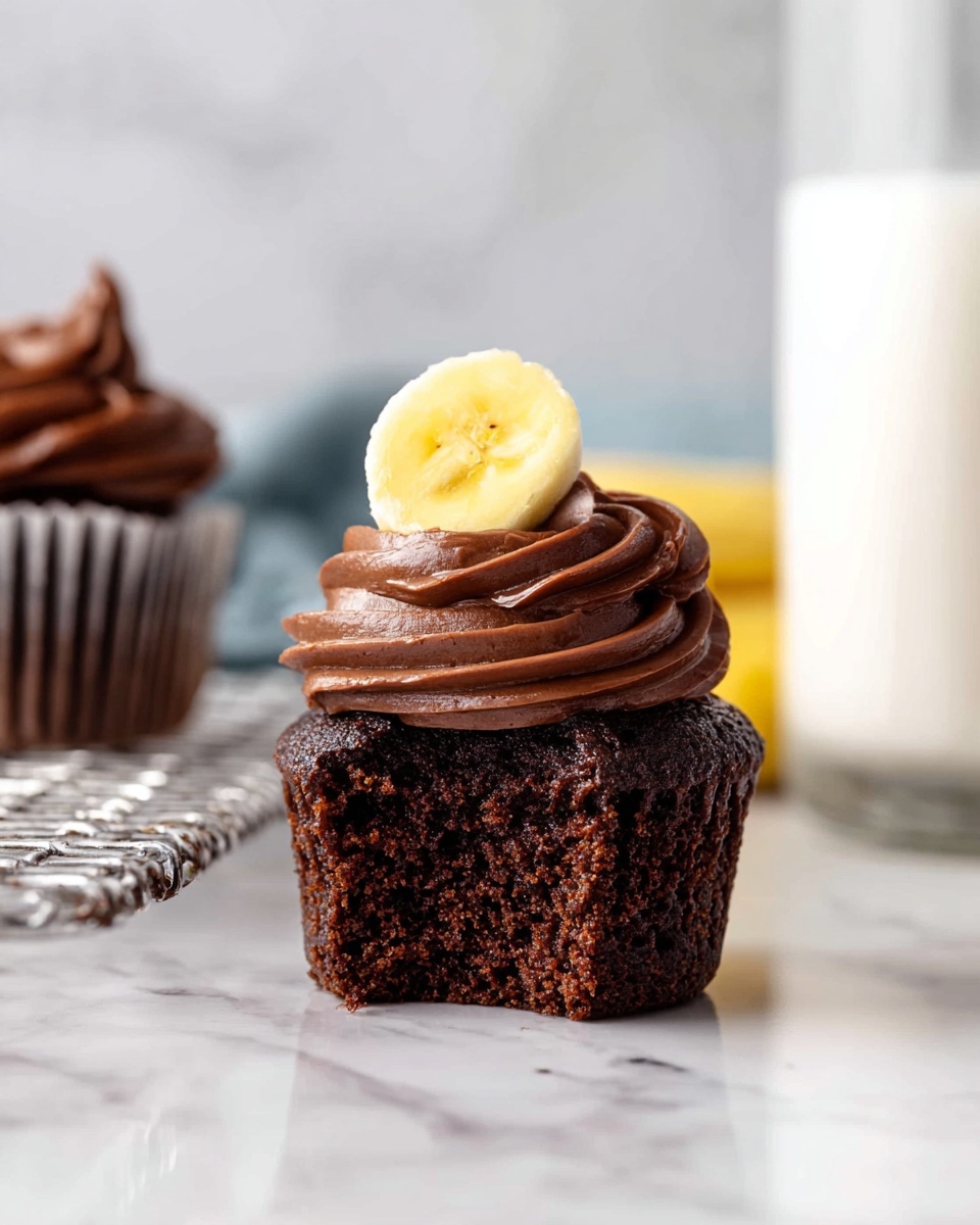 The image shows a chocolate cupcake with three layers: the bottom layer is rich, dark brown chocolate cake with a moist texture, the middle layer is a thick swirl of smooth, dark chocolate frosting covering the top of the cake, and the top layer is a single crisp yellow banana chip placed on the edge of the frosting. In the background, there is another cupcake with similar layers cooling on a white metal rack. The surface under the cupcake is a shiny white marble texture. A clear glass of milk is partially visible on the right side, adding to the scene. Photo taken with an iphone --ar 4:5 --v 7