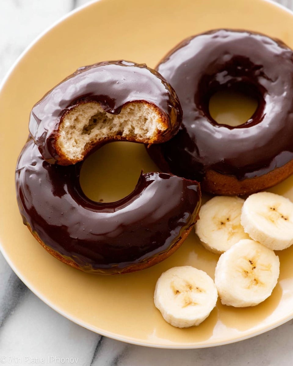 Two chocolate glazed donuts sit on a white plate with a soft yellow tone. One donut is whole, showing a smooth and shiny dark chocolate glaze covering its round shape. The other donut is broken in half, revealing a light brown, fluffy inside and a thick layer of the same dark chocolate glaze on top. Nearby, three slices of banana rest on the plate, adding a fresh, light cream color to the scene. The plate is placed on a white marbled surface. photo taken with an iphone --ar 4:5 --v 7