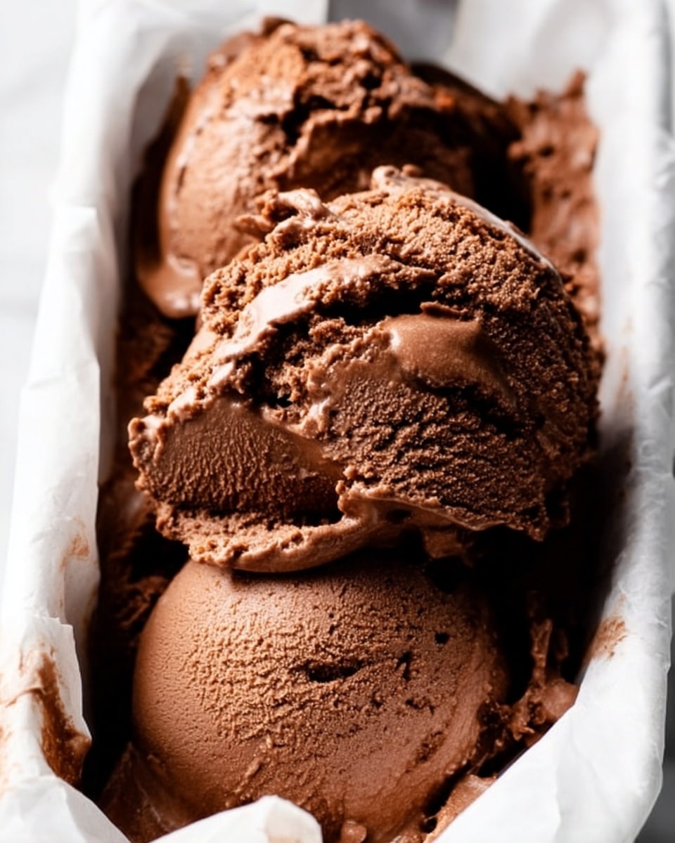 The image shows a close-up of rich brown chocolate ice cream scooped into a white container with slight crinkles of white paper around it. The ice cream has a creamy and smooth texture with visible small swirls and chunks, giving it a thick and dense look. The light highlights the slight shine and airiness of the scoops, which sit one on top of the other in thick layers. The background features a white marbled texture. photo taken with an iphone --ar 4:5 --v 7