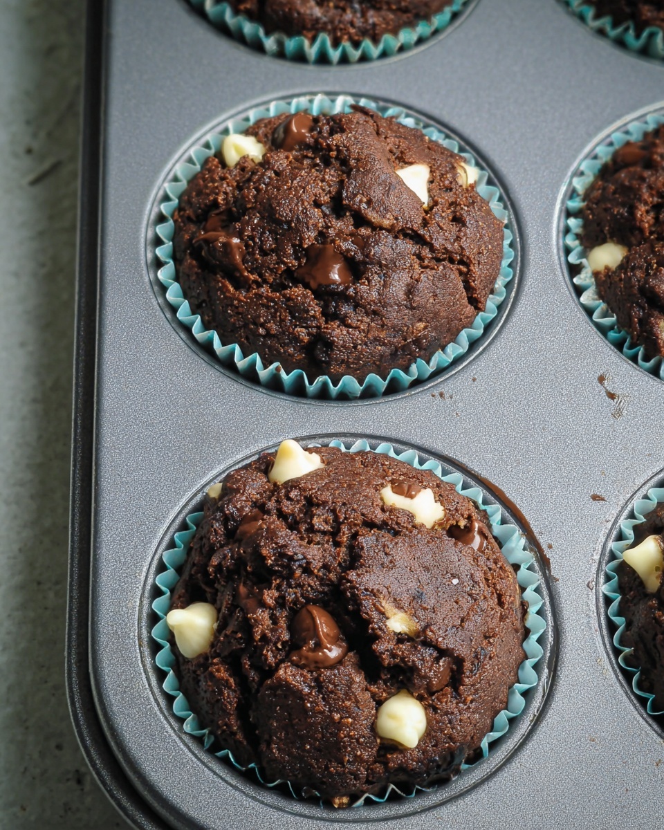 The image shows three dark brown chocolate muffins with a rough, cracked top surface nestled in light blue paper liners. Each muffin is studded with unevenly scattered white and dark chocolate chips that slightly melt into the warm, chewy texture. The muffins sit inside a gray metal muffin tray, with the tray placed on a white marbled surface partly visible at the edges. The muffins have a rich, dense look with a few small air pockets and a glossy finish on some chocolate chips. photo taken with an iphone --ar 4:5 --v 7