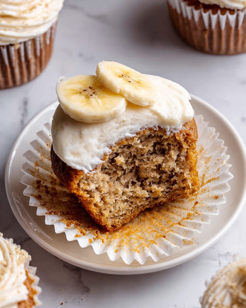 A half-eaten banana cupcake sits on a white plate with a white cupcake liner underneath. The cupcake has a moist, textured light brown cake layer visible inside. On top, there is a layer of smooth white cream frosting, slightly swirled, with two thin banana slices placed neatly on one side near the frosting. The plate rests on a white marbled surface with parts of other similarly decorated cupcakes partially visible around the edges. photo taken with an iphone --ar 4:5 --v 7