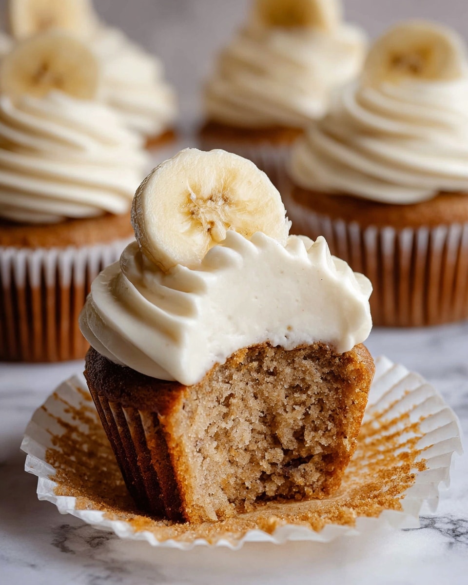 The image shows a cupcake with three clear layers. The bottom layer is a light brown, moist cake with small pieces giving it a textured look. On top is a thick, swirled layer of creamy white frosting, soft and smooth in texture. The topmost layer is a thin, round banana slice placed vertically at the center of the frosting. The cupcake is in a white paper liner that is partly peeled back to show the cake inside. Surrounding it are similar cupcakes, each with the same three-layer look and a banana slice on top, all sitting on a white marbled surface. Photo taken with an iphone --ar 4:5 --v 7
