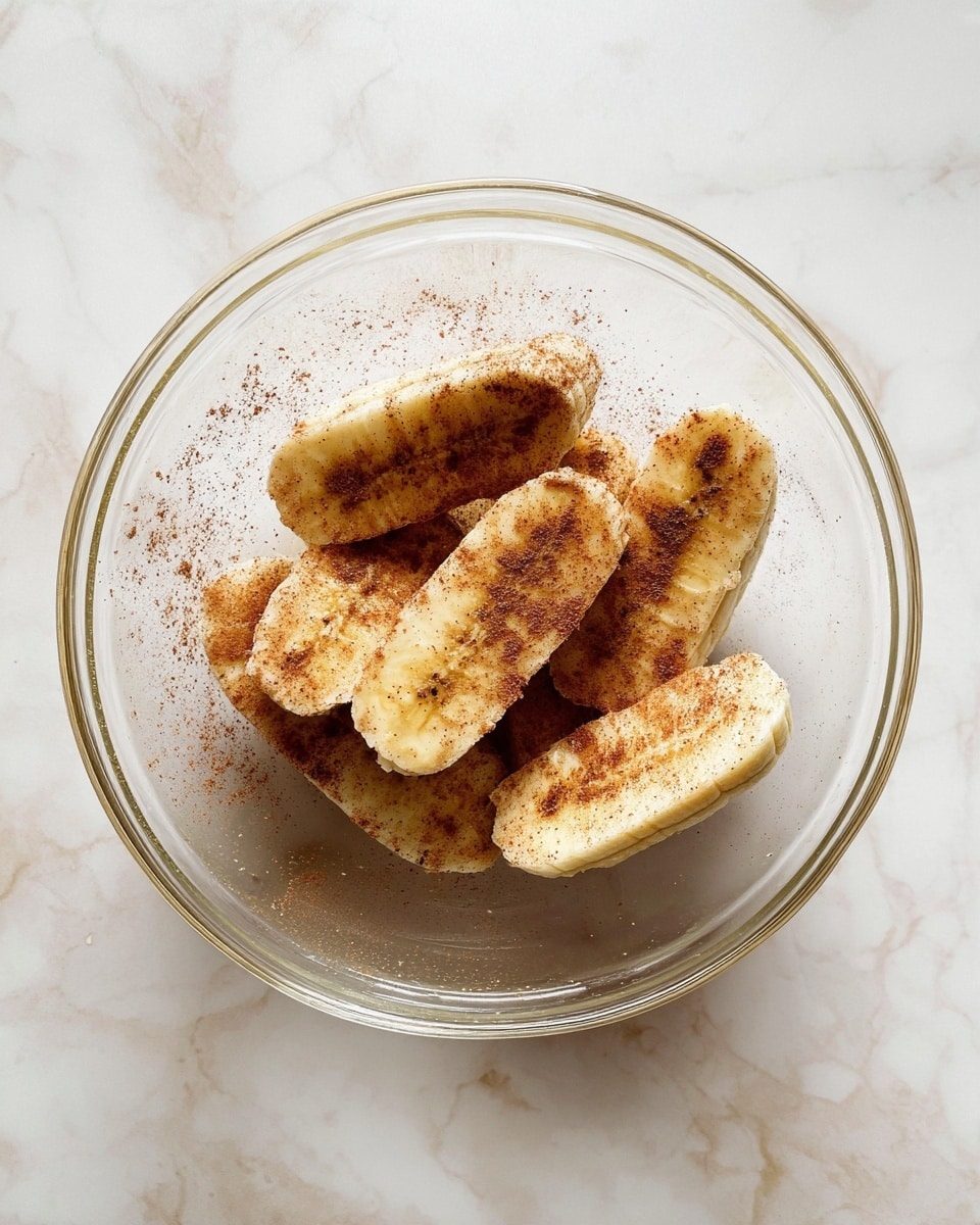 A clear glass bowl on a white marbled surface holds seven banana halves, each peeled and coated with a light layer of brown cinnamon powder. The bananas are arranged in a loose pile, showing their pale yellow color with specks of cinnamon evenly spread on them. The inside of the glass bowl shows faint smudges of cinnamon powder, enhancing the natural texture of the bananas. The overall image has soft natural light, making the bananas look fresh and warm. photo taken with an iphone --ar 4:5 --v 7