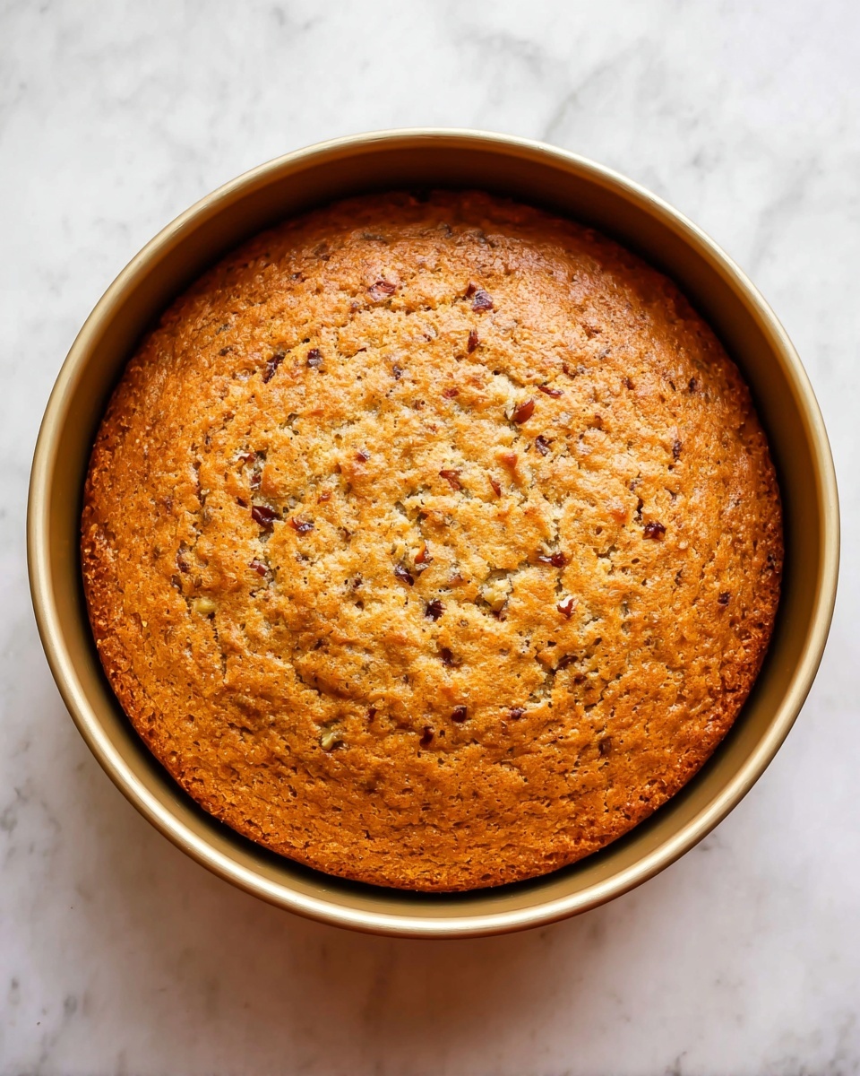 A round golden brown cake sits inside a light gold baking pan, filling it fully with a slightly uneven top texture showing small bits of nuts and possibly fruit mixed in, giving it a speckled look of darker and lighter brown spots. The surface looks soft with a few small cracks and the pan is placed on a white marbled surface. photo taken with an iphone --ar 4:5 --v 7