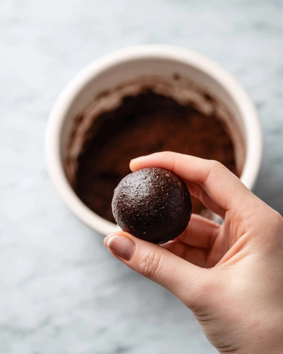 A close-up of a woman's hand holding a small, dark brown, smooth ball of dough or mixture, shaped roughly round with a slight shine. In the background, there is a white pot filled with more of the same dark brown mixture, blurred to keep focus on the ball in the woman's hand. The scene is set against a white marbled surface. photo taken with an iphone --ar 4:5 --v 7