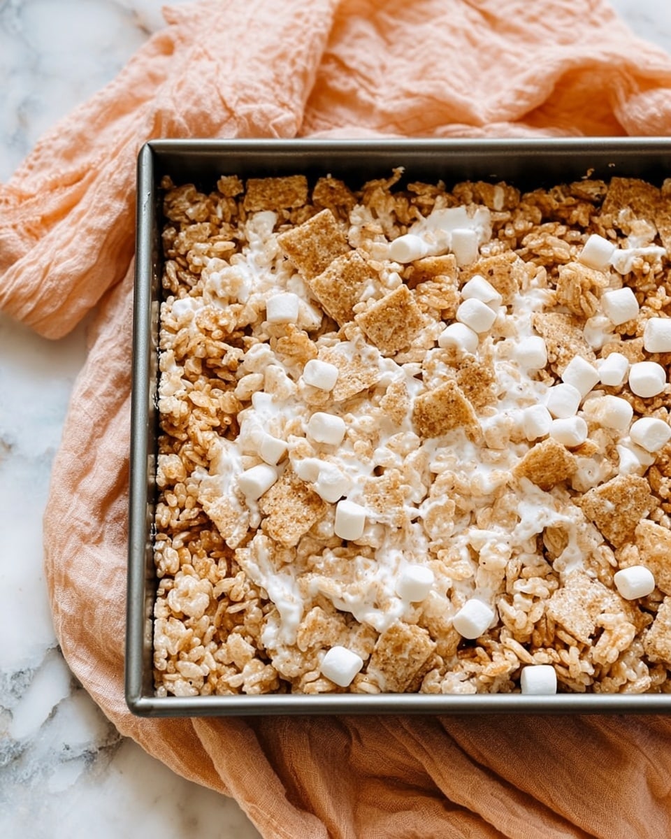 A close-up view of a square pan filled with a crispy layered dessert made of puffed rice cereal and golden brown cereal squares, mixed with soft, melted white marshmallows that create stringy textures across the top. The dessert is dense, with a mix of light beige and pale brown pieces, and scattered small white marshmallows are visible throughout. The pan sits on a white marbled surface with a soft, crinkled peach cloth partially underneath it. Photo taken with an iphone --ar 4:5 --v 7