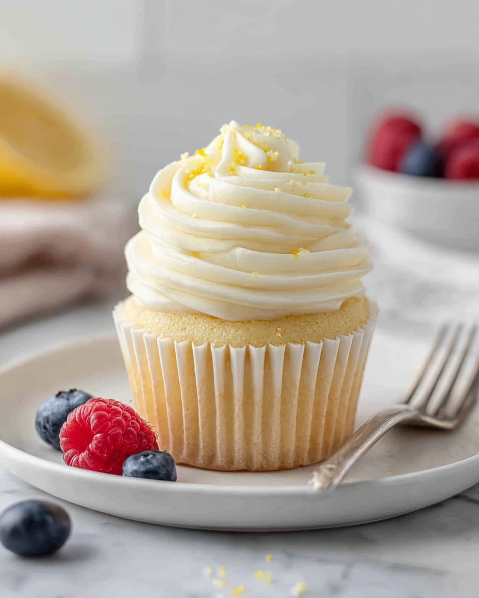 A vanilla cupcake sits in the center of a white plate on a white marbled surface. The cupcake has one layer of soft yellow cake wrapped in a ridged light beige paper liner. On top is a thick swirl of creamy white frosting that spirals upward in three smooth, rounded layers, sprinkled lightly with tiny yellow bits. Around the plate are a few scattered fresh blueberries and raspberries, adding pops of deep blue and bright red color. A silver fork rests partly on the plate to the right, and soft natural light brightens the scene. photo taken with an iphone --ar 4:5 --v 7