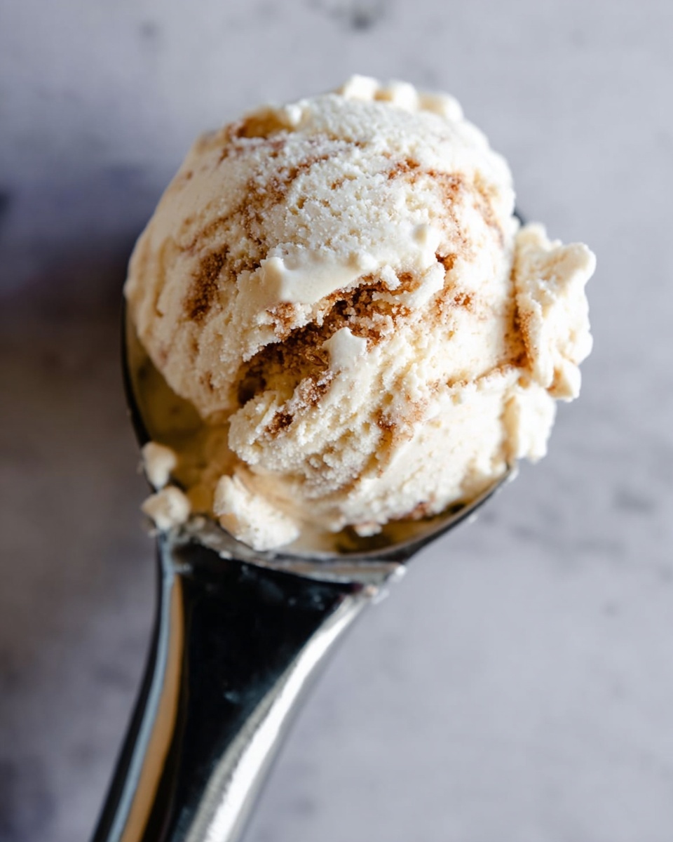 The image shows a close-up of a single scoop of ice cream held by a metal ice cream scooper. The ice cream scoop is creamy white with visible brown swirls mixed throughout, creating a textured look. The background is a smooth white marbled surface that contrasts with the metal scooper’s shiny silver color. The scoop is round and slightly uneven with some ice cream clinging to the edges of the scooper. Photo taken with an iphone --ar 4:5 --v 7