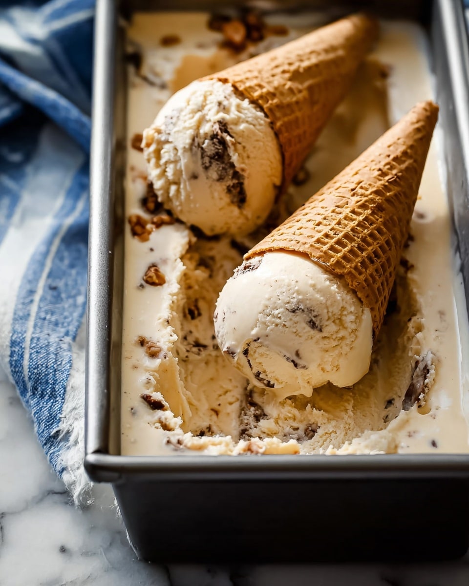 The image shows a rectangular metallic pan filled with light beige ice cream that has dark brown swirls and chunks spread throughout. Two scoops of this ice cream are placed on top of the surface in the pan, each scoop topped with a light brown waffle cone tilted slightly to the side. The texture of the ice cream looks creamy and smooth with visible bits embedded inside. The background features a white marbled surface with a cloth that has blue and white stripes partially visible under the pan. The overall look is rich and inviting. photo taken with an iphone --ar 4:5 --v 7