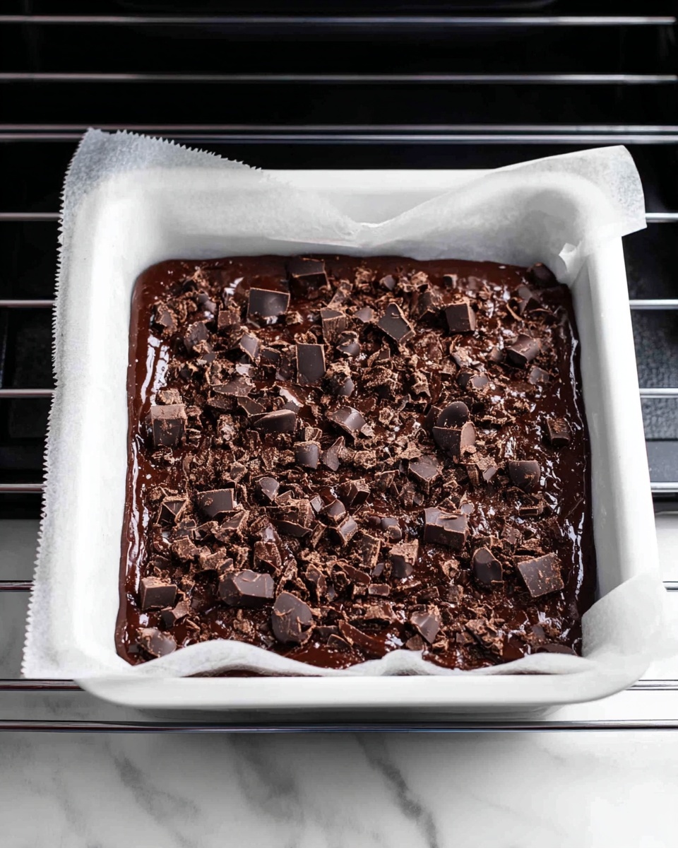 A white baking tray lined with white parchment paper holds a single thick layer of dark brown chocolate batter spread evenly inside. The batter is topped with a generous layer of small, uneven chocolate chunks scattered all over the surface. The tray is set on a black oven rack with a white marbled texture visible beneath. The edges of the parchment paper slightly rise above the tray, framing the rich chocolate mixture. Photo taken with an iphone --ar 4:5 --v 7