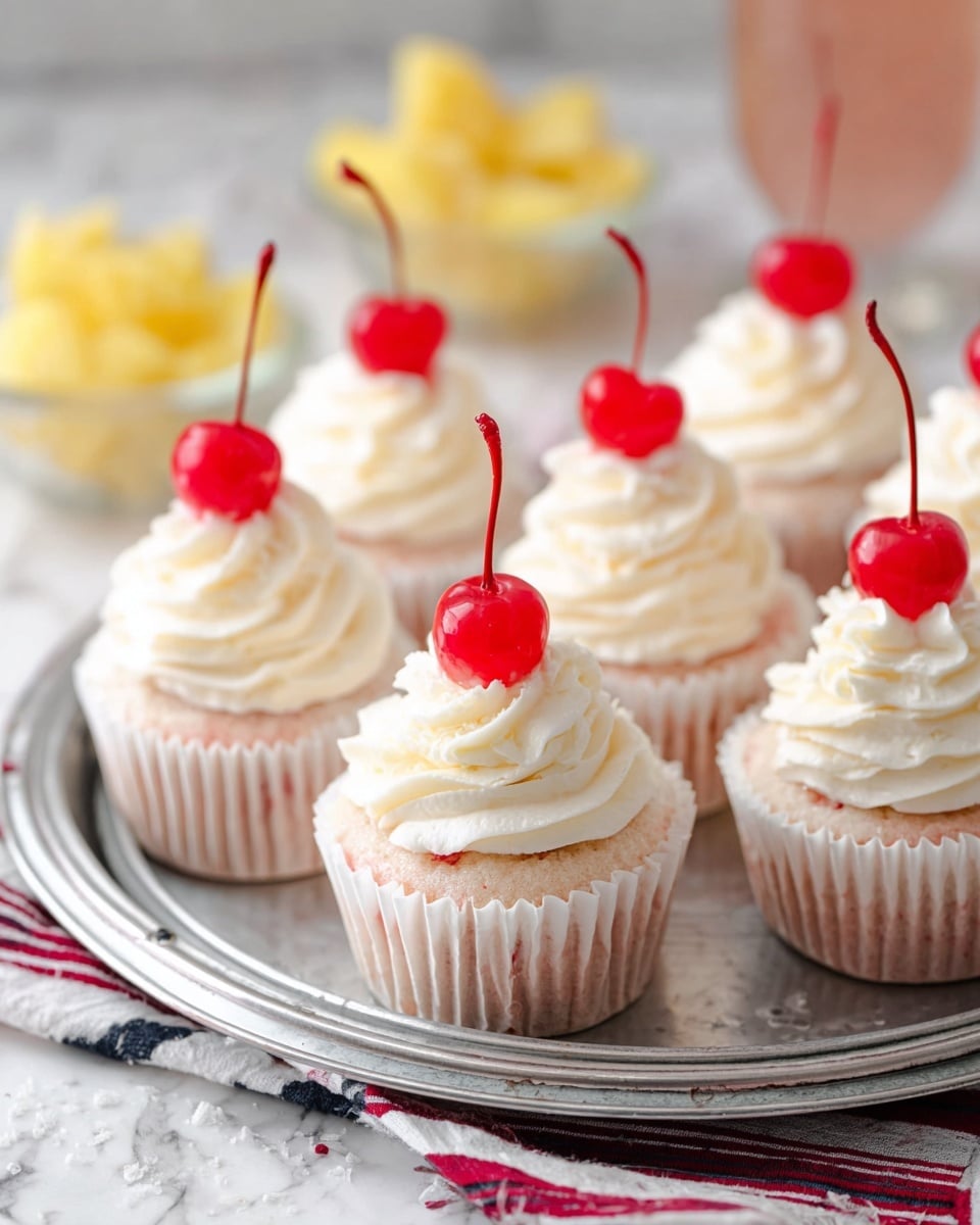 The image shows a round metal tray filled with small cupcakes, each wrapped in white paper liners. Each cupcake has two main layers: a dense light pink cake base and a swirl of smooth white frosting on top. On top of the frosting sits a bright red cherry with a long thin stem. The tray is placed on a cloth with red, beige, and dark blue stripes, all set on a white marbled surface. In the background, slightly out of focus, there are small bowls holding yellow pineapple chunks and more red cherries. The lighting is soft, making the colors pop gently. photo taken with an iphone --ar 4:5 --v 7