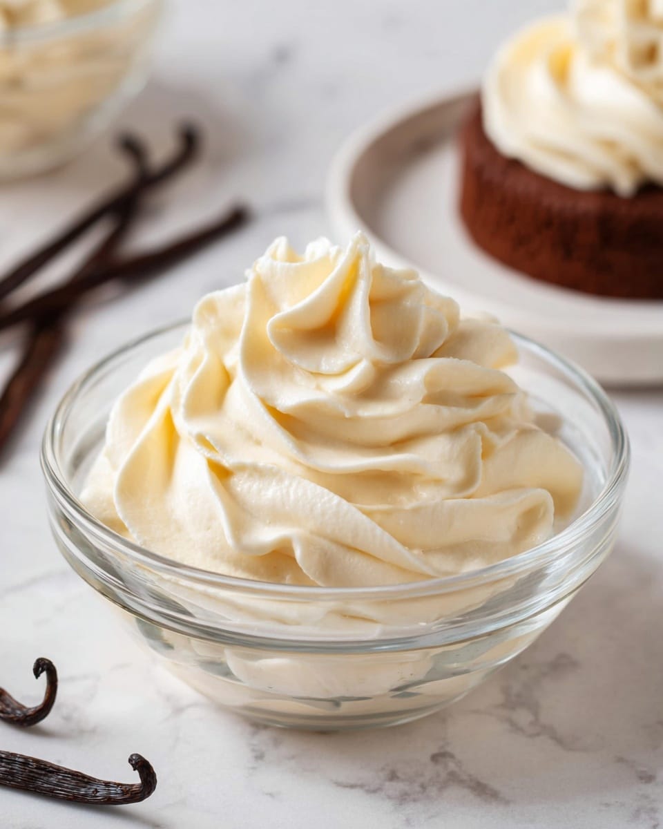 The image shows a close-up of a transparent glass bowl filled with light creamy beige whipped frosting or mousse, shaped into smooth, soft, and thick swirls with elegant peaks and folds. The bowl sits on a white marbled surface. In the background, there is a blurred white plate holding a small chocolate cake topped with a similar creamy swirl. To the left of the bowl, dark brown vanilla pods, slightly curved, rest on the surface, adding contrast. The overall texture of the cream looks smooth and airy, with soft shadows and highlights that capture its lightness. Photo taken with an iphone --ar 4:5 --v 7
