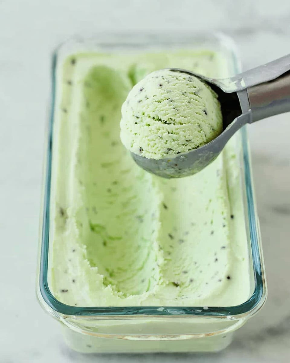 A clear glass rectangular container holds light green ice cream with small dark green specks throughout, showing a smooth and creamy texture. A metal ice cream scoop lifts a round, full scoop of the same green ice cream above the container, capturing the soft, dense look. The background features a white marbled surface. photo taken with an iphone --ar 4:5 --v 7