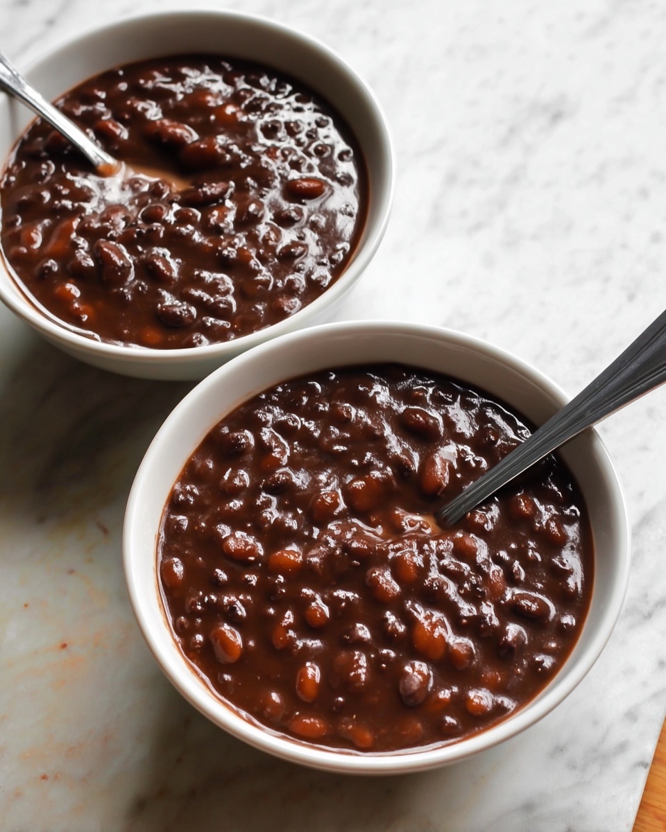 Two white bowls filled with thick, dark brown bean stew with visible soft beans throughout. Each bowl has a silver spoon partially inside, one spoon angled toward the camera. The bowls sit on a white marbled textured surface, creating a clean and bright background. The stew looks shiny and rich, with a smooth texture combined with tender bean pieces. photo taken with an iphone --ar 4:5 --v 7