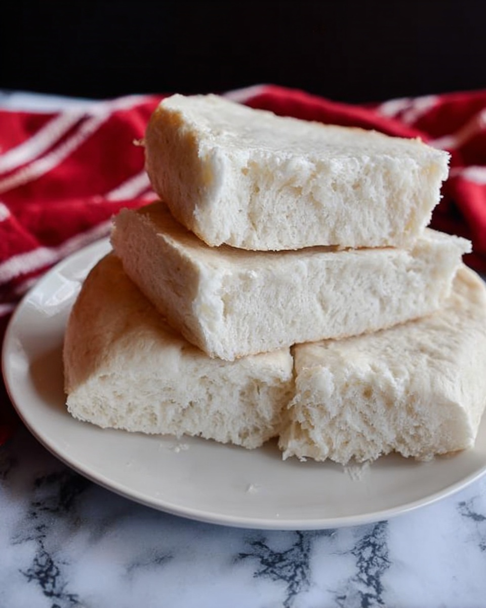 The image shows four pieces of soft, white bread with a slightly crumbly texture, stacked in layers on a white plate. The top piece is tilted to reveal its fluffy inside. The plate is placed on a white marbled surface with a red and white cloth in the background. The bread looks fresh and homespun, with no extra toppings or decorations. photo taken with an iphone --ar 4:5 --v 7