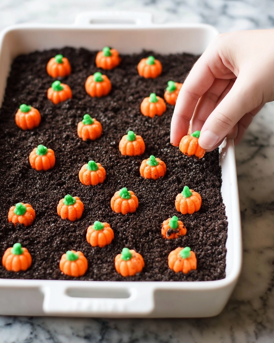 The image shows a white rectangular dish filled with a thick, dark brown crumbly layer that looks like dirt on the surface. On top of the dark base, there are many small orange candy pumpkins with green tops scattered evenly across the dish. A woman's hand is placing one of these small candy pumpkins onto the crumbly dark layer. The background is a white marbled texture. photo taken with an iphone --ar 4:5 --v 7