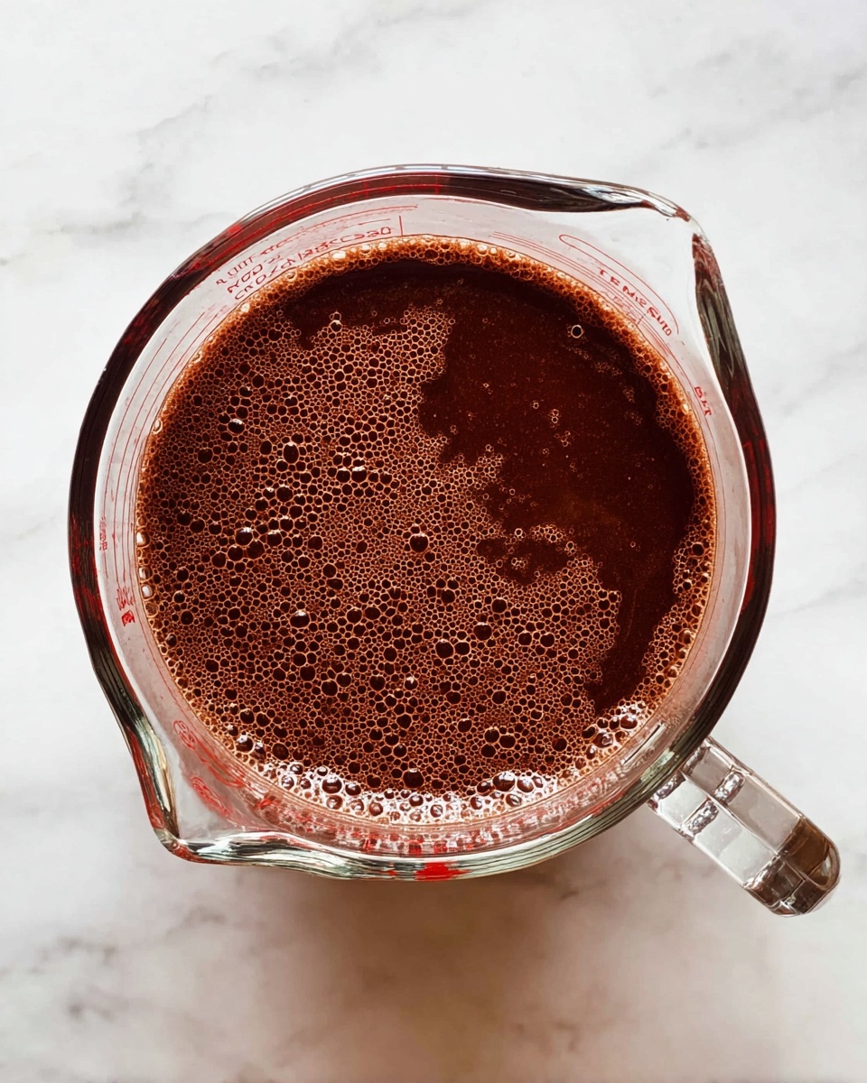 A top-down view of a clear glass measuring cup filled with a thick, dark brown liquid that has many small bubbles on its surface. The measuring cup has a clear handle on the right side and is placed on a white marbled surface, which contrasts with the dark liquid inside. The texture of the liquid looks smooth and slightly frothy, filling the cup almost to the brim. photo taken with an iphone --ar 4:5 --v 7