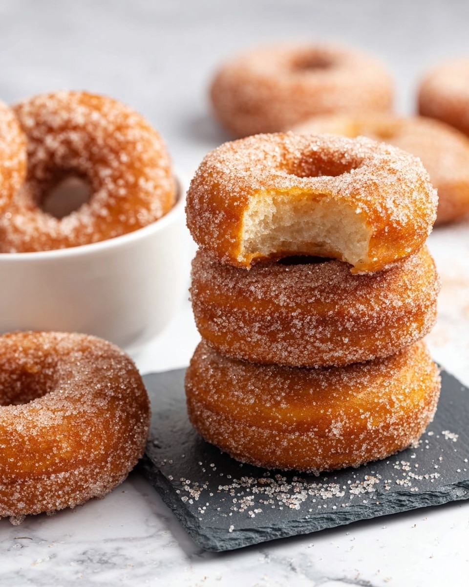 There is a close-up of a stack of three round doughnuts covered in sugar crystals on a dark slate square, with the top doughnut showing a bite taken out to reveal a soft, light inside. To the side, a white bowl holds more sugar-coated doughnuts, and several more are scattered lightly blurred on a white marbled surface in the background. The doughnuts have a golden brown color with a rough sugar texture evenly coating their outside, and the overall scene is bright and clean. Photo taken with an iphone --ar 4:5 --v 7