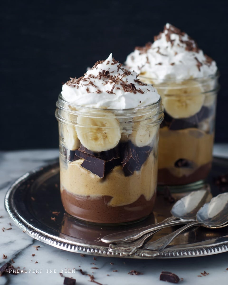 Two clear glass jars filled with layered dessert sit on a silver tray with two silver spoons next to them. Each jar has three layers: the bottom layer is dark chocolate pieces, the middle layer is light brown creamy pudding with banana slices pressed against the glass, and the top layer is whipped white cream sprinkled with small chocolate shavings. The background is dark, with a white marbled texture surface. photo taken with an iphone --ar 4:5 --v 7
