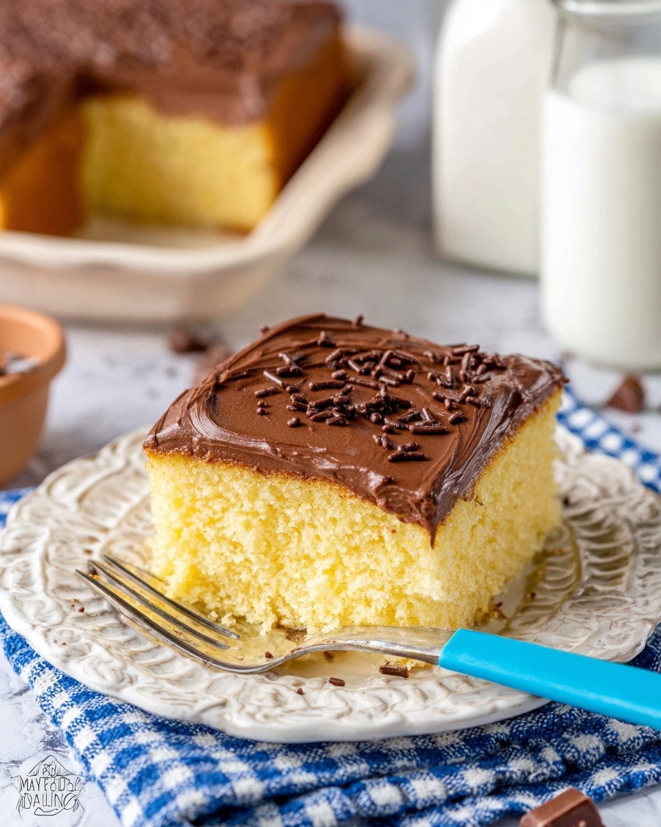 A square piece of yellow cake sits on a white decorative plate, topped with a thick layer of smooth chocolate frosting. The chocolate frosting is spread evenly over the top with scattered chocolate sprinkles. The cake has a soft and fluffy texture with one slice slightly lifted by a fork with a blue handle, showing the pale yellow inside. The scene is set on a white marbled textured surface with a blue and white checkered cloth nearby and blurred baking items in the background. Photo taken with an iphone --ar 4:5 --v 7