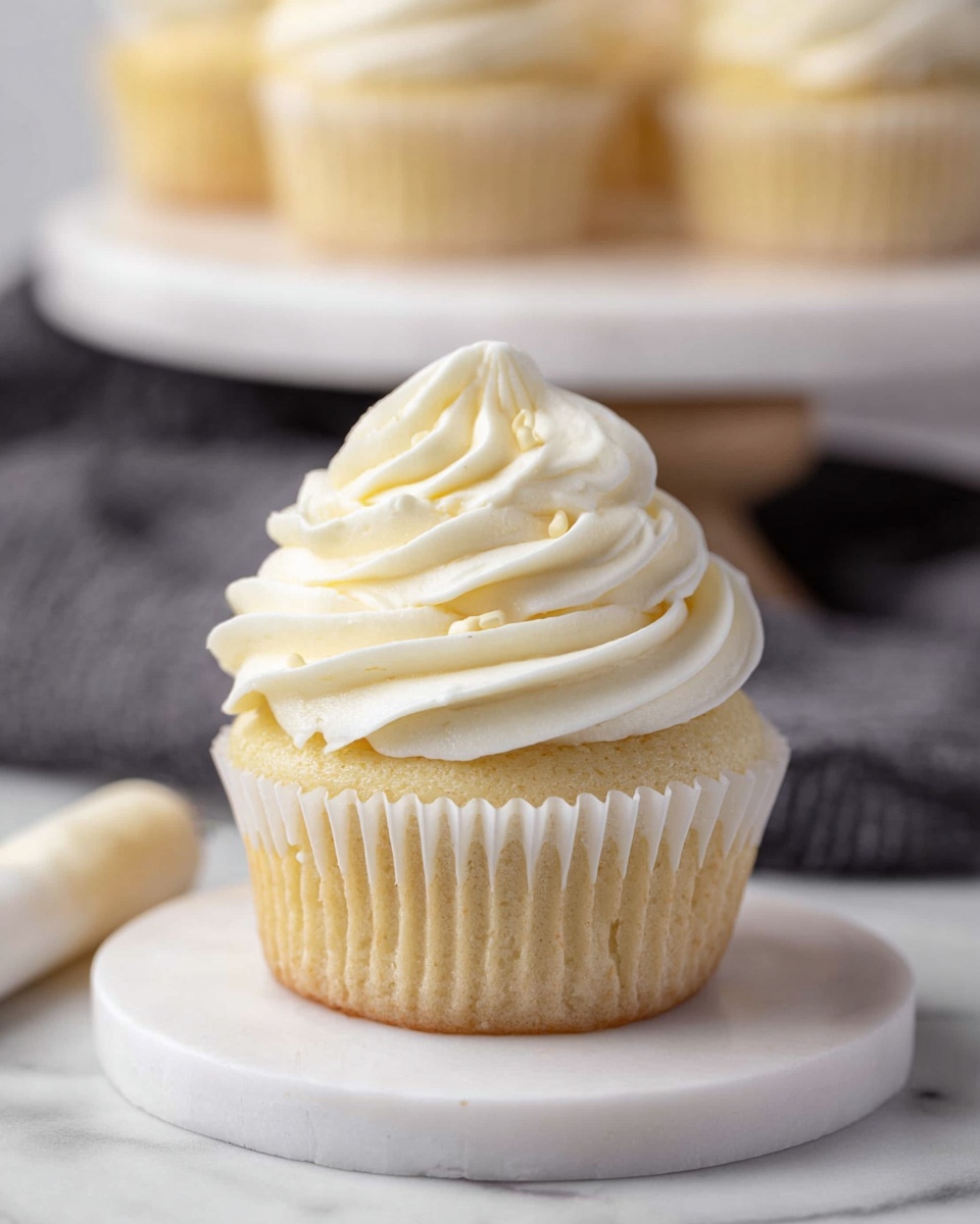 Two light yellow cupcakes sit on a wooden board placed on a white marbled surface. Each cupcake has a thick swirl of creamy white frosting on top with a smooth, soft texture and distinct ridges, forming a tall, peaked shape. The cupcake liners are slightly translucent, showing the soft cake beneath. In the background, there is a blurred white bowl filled with more of the same white frosting, adding depth to the image. The lighting is soft and even, highlighting the details in the frosting ridges and the texture of the cake. photo taken with an iphone --ar 4:5 --v 7