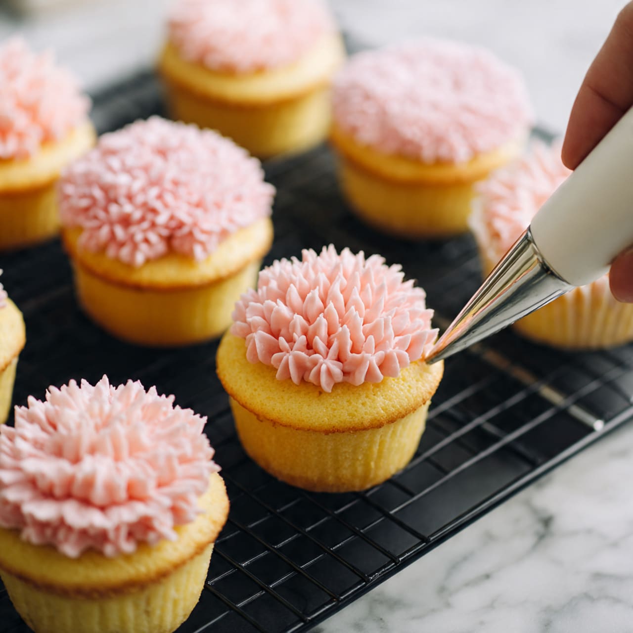 The image shows several yellow cupcakes on a black cooling rack with a white marbled surface underneath. Each cupcake has a soft, smooth yellow layer in the center, which looks like custard or cream. Some cupcakes are fully topped with light pink frosting shaped into many small, soft, wavy petals, creating a fluffy flower-like texture that covers the whole top. One cupcake has a half decoration of these pink petals on one side, revealing the smooth yellow center on the other half. A white piping bag is visible in the lower right corner with a silver tip, indicating the frosting is being applied. Photo taken with an iphone --ar 4:5 --v 7