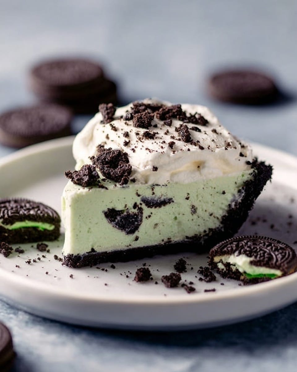 A slice of pie sits centered on a white plate with a smooth edge, placed on a white marbled textured surface. The bottom layer of the pie is a dark chocolate crumb crust, thick and firm. Above the crust is a creamy, light green mint-flavored filling scattered with small, dark cookie pieces throughout. The top layer is a thick, fluffy white whipped cream dollop that covers the pie slice partially, with more crushed cookie crumbs lightly sprinkled on top. There are some dark chocolate mint sandwich cookies placed around the plate, some whole and one broken to show the green filling inside. Photo taken with an iphone --ar 4:5 --v 7