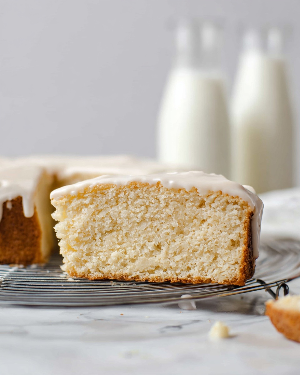 The image shows a single slice of a thick, soft cake with a light beige color and crumbly texture. The top layer has a smooth white glaze that slightly drips down the side. In the foreground, there is a small piece of the cake also topped with the same white glaze. The cake sits on a metal rack on a white marbled surface. In the blurred background, there are two glass bottles filled with milk. Photo taken with an iphone --ar 4:5 --v 7