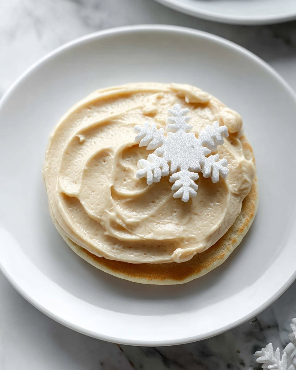 A single light brown pancake sits on a white plate with a smooth, creamy layer of light beige frosting spread evenly on top, showing slight swirling textures. On the frosting, there is a white snowflake-shaped decoration placed near the edge. The plate is set on a white marbled surface. Photo taken with an iphone --ar 4:5 --v 7