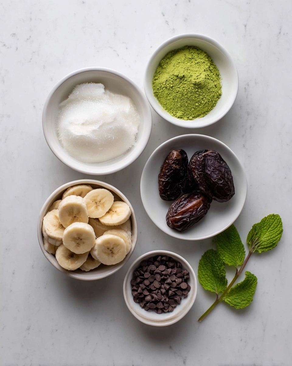 The image shows five white bowls of different sizes arranged on a white marbled surface with a small sprig of fresh green mint leaves to the right. The largest bowl at the bottom left is filled with light brown frozen banana slices. Above it to the left, a medium bowl contains smooth white coconut oil. To the right of the coconut oil, a small bowl holds bright green matcha powder on top of a white base. Below the matcha powder, a small bowl contains dark brown dried dates. At the bottom right, the smallest bowl is filled with tiny dark brown chocolate chips. All bowls have a smooth, clean texture, and the whole composition is neat and simple with soft natural light. Photo taken with an iphone --ar 4:5 --v 7