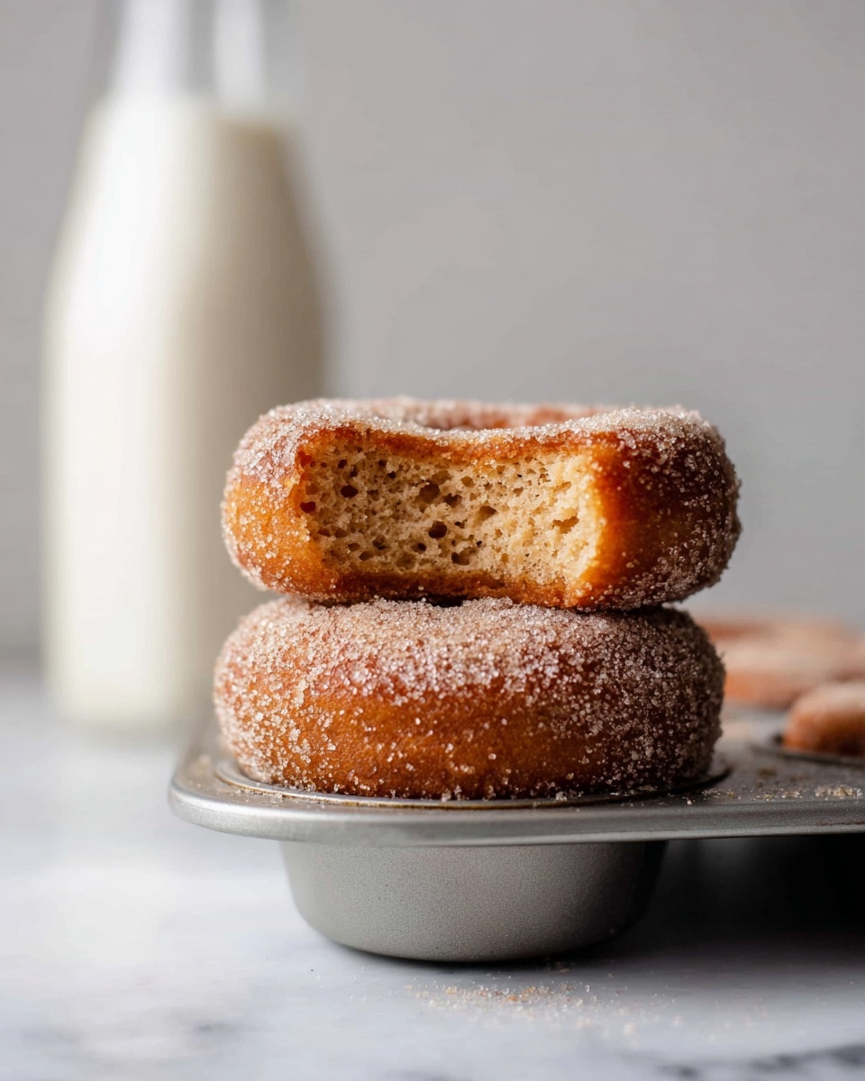 Two soft, round donuts stacked on top of each other inside a silver metal muffin pan, each donut covered in a sparkling layer of sugar grains giving a rough texture; the top donut has a bite taken out, showing a light brown, fluffy inside with small crumb holes. In the background, there is a tall, blurry white bottle standing upright on a white marbled surface. photo taken with an iphone --ar 4:5 --v 7