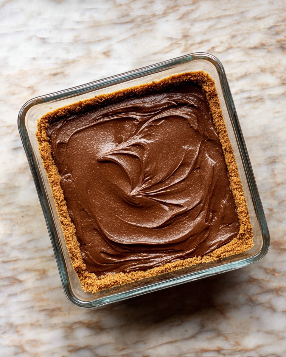 A clear square glass dish contains a two-layer dessert placed on a white marbled textured surface. The bottom layer is a coarse, light brown crumb crust pressed evenly along the base and edges of the dish. The top layer is a smooth, rich dark brown chocolate-like filling spread thickly and evenly with soft swirls visible across the surface. The layers contrast in texture with the crumbly base and creamy top. Photo taken with an iphone --ar 4:5 --v 7