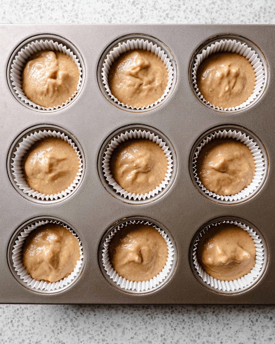 The image shows nine white paper cupcake liners filled with light brown smooth batter, placed evenly in a metallic cupcake tray. Each liner is about three-quarters full with soft, slightly textured batter that has small air bubbles and gentle peaks on the surface. The tray’s metal surface contrasts with the warm batter and crisp white liners. The background is a white marbled texture. photo taken with an iphone --ar 4:5 --v 7