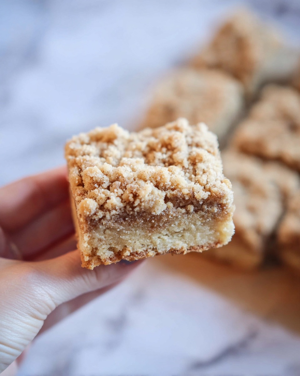 A close-up image shows a woman's hand holding a single square crumb cake piece. The cake has two visible layers: a light tan base layer with a soft texture and small visible pieces inside, and a thick crumb topping with a slightly darker tan color made of small, coarse crumbs. The crumb layer looks rough and crumbly, covering the entire top. The background shows more pieces of the same crumb cake on a white marbled surface, softly blurred to keep focus on the held piece. Photo taken with an iphone --ar 4:5 --v 7