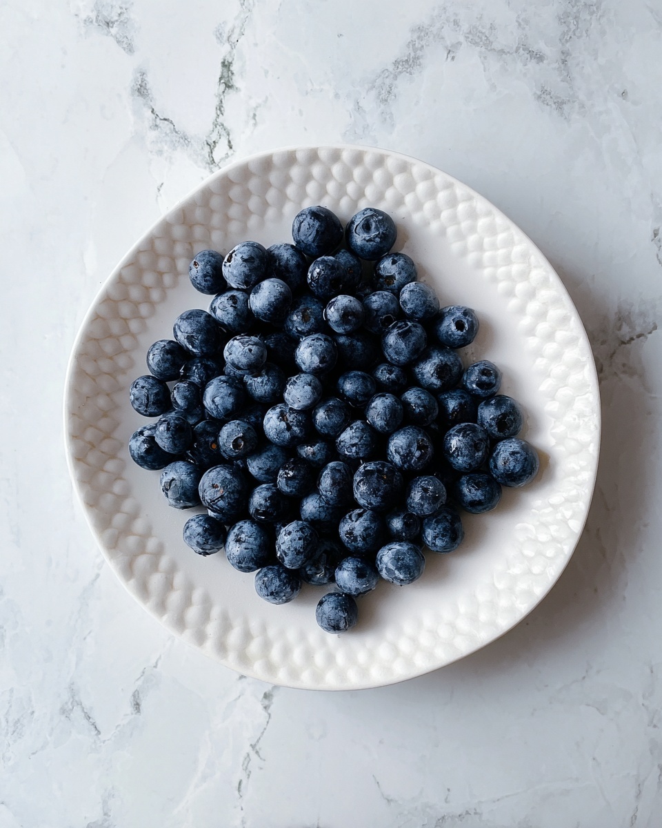 A white plate with small rounded edges holds one layer of fresh blueberries, deep blue with a slight frost-like texture on their skin. The plate sits on a white marbled surface with soft gray veining, creating a clean and simple background. photo taken with an iphone --ar 4:5 --v 7