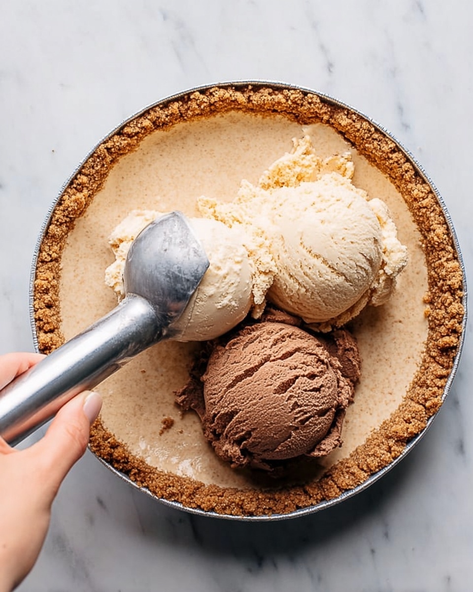 A close-up image of a pie tin with a light brown crumb crust inside, two scoops of ice cream placed on the crust. One scoop is light beige, and the other is chocolate brown. A woman's hand is holding a metal ice cream scooper that is partly under the beige scoop. The tin rests on a white marbled surface photo taken with an iphone --ar 4:5 --v 7