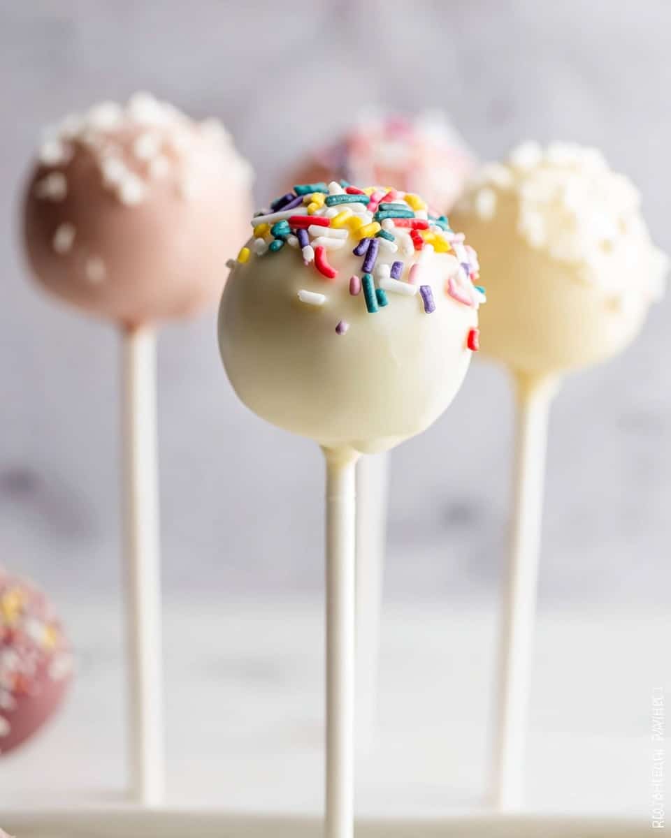 The image shows a close-up of four cake pops on white sticks, standing upright on a white surface with a white marbled background. The nearest cake pop is coated in smooth white icing and decorated on top with colorful sprinkles in red, yellow, green, blue, white, and purple. Behind it, there are three other cake pops with a similar smooth finish in light pastel colors, including pale pink and cream, some topped with tiny white sprinkles. The overall look is delicate and clean, with soft lighting highlighting the creamy texture of the cake pop coating. photo taken with an iphone --ar 4:5 --v 7