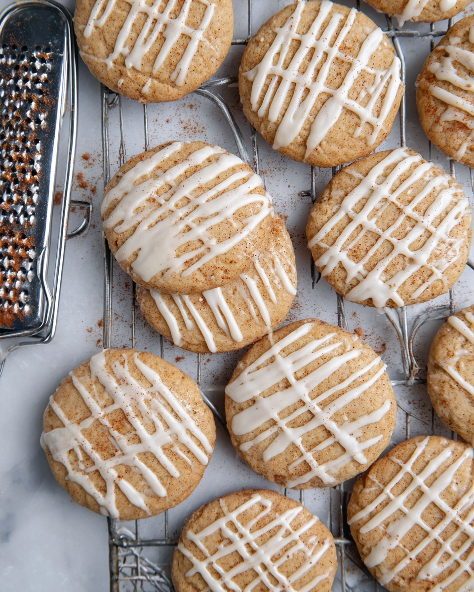 A group of round cookies with a light brown color is placed on a metal cooling rack. Each cookie has a thick drizzle of white icing creating a grid-like pattern on top. The cookies appear soft with slightly uneven edges and some light specks of brown, possibly cinnamon, sprinkled over the icing. To the left side, there is a metal grater partially shown, with some brown spice on it. The whole scene is set on a white marbled surface. photo taken with an iphone --ar 4:5 --v 7