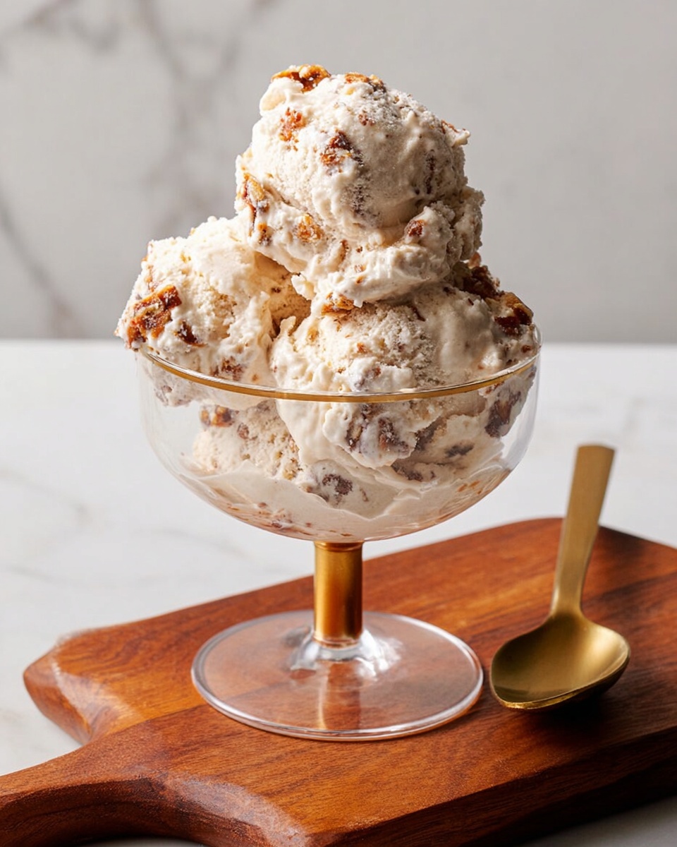 A clear glass dessert cup with a gold base holds three scoops of creamy ice cream mixed with small brown and lighter beige bits, showing a rough, chunky texture. The ice cream scoops are stacked to form one layer at the bottom and two on top, slightly uneven with some soft melting at the edges. The cup is placed on a warm brown wooden board, with a gold spoon lying next to it. The background is a clean white marbled surface. Photo taken with an iphone --ar 4:5 --v 7