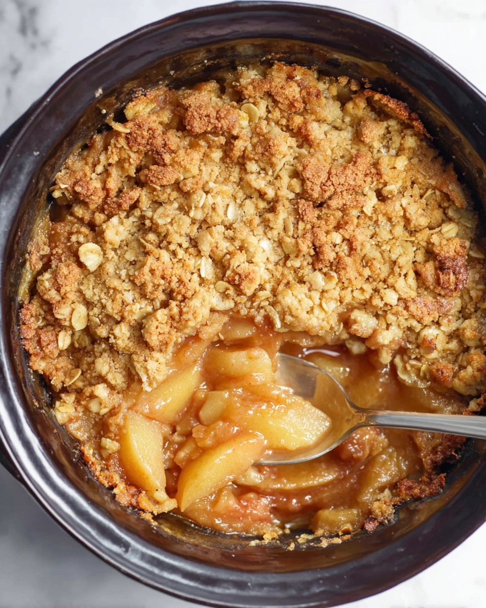 A round dish shows a baked apple crumble inside a dark cooking pot. The top layer is a golden, crumbly oat topping with coarse chunks, showing varying browns and light tan colors. Beneath this, a thick, glossy layer of soft, warm apple slices in syrupy caramel color is partially visible where a silver spoon scoops out some filling. The apple slices are light yellow and cover the bottom layer inside the pot. The pot rests on a white marbled surface. photo taken with an iphone --ar 4:5 --v 7