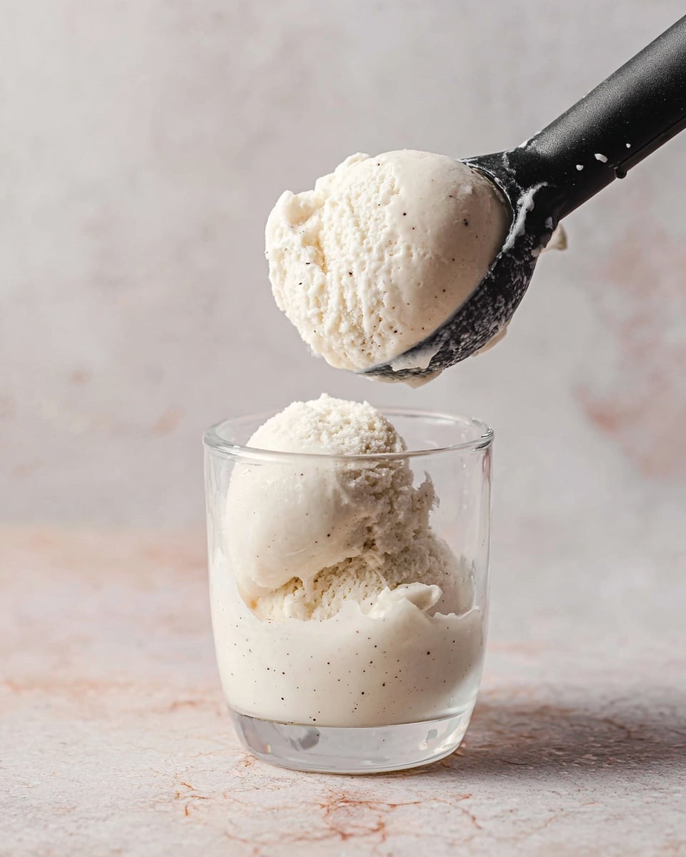 A clear glass cup holds one scoop of creamy white ice cream with tiny dark specks, showing a smooth and slightly soft texture. Above it, a black ice cream scooper lifts a second scoop of the same ice cream, highlighting its thick, cold consistency, with faint specks visible throughout. The scene is set on a surface with a white marbled texture and a soft, blurry background. photo taken with an iphone --ar 4:5 --v 7