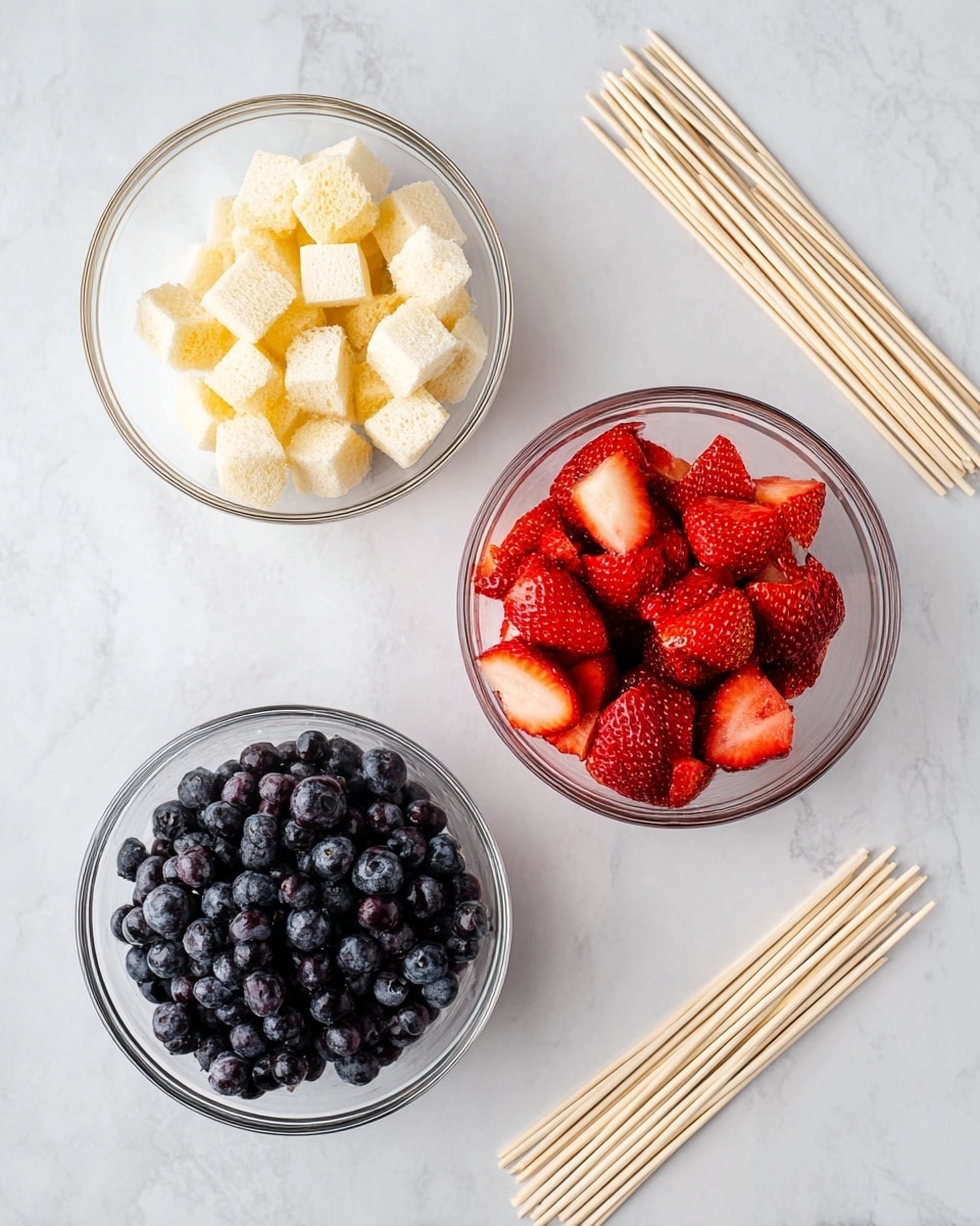Three clear glass bowls sit on a white marbled surface. The top left bowl holds small, light yellow cubes of bread with a soft texture. The top right bowl is filled with bright red strawberries, some whole and some cut in half, showing their juicy, seeded inside. The bottom left bowl contains dark blue blueberries with a smooth, round shape. To the far right, a bunch of light wooden skewers are laid flat on the white marbled surface. Photo taken with an iphone --ar 4:5 --v 7