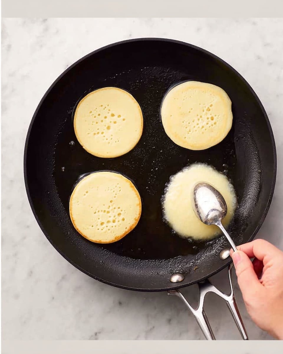 The image shows a black pan on a white marbled surface with three small, round pancake batter blobs cooking. A woman's hand holds a silver spoon, scooping some batter onto the pan. The pancakes are pale yellow and smooth, with one pancake having small bubbles on its surface, indicating cooking progress. The overall scene captures the process of making pancakes. Photo taken with an iphone --ar 4:5 --v 7